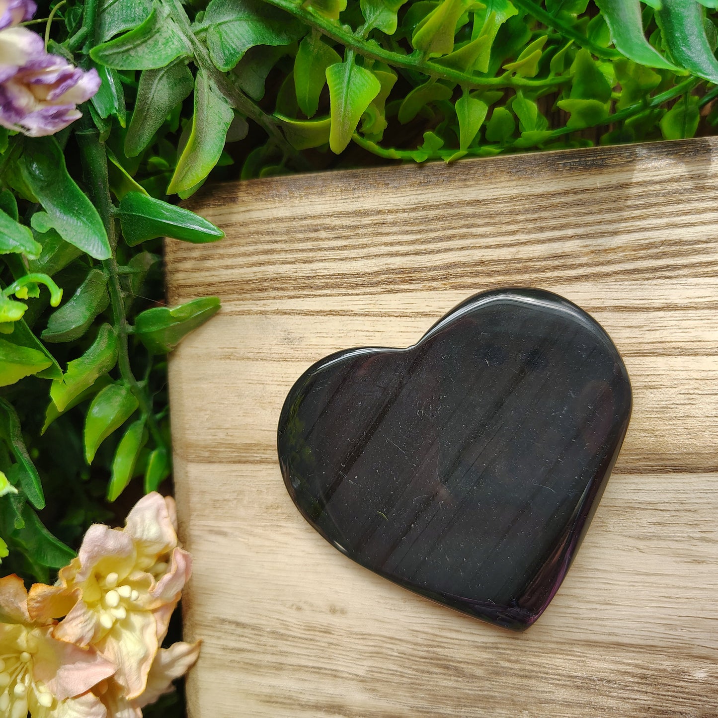 Heart-shaped black stone on a wooden surface with greenery and flowers in the background