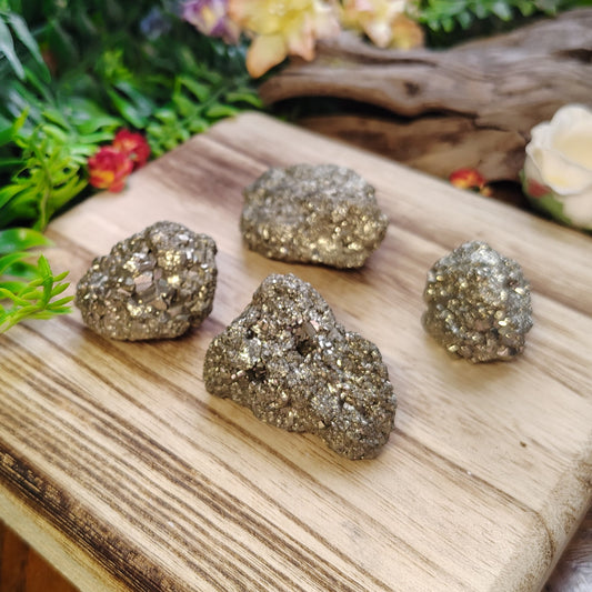 Four metallic pyrite crystals on a wooden board with decorative plants and flowers in the background.