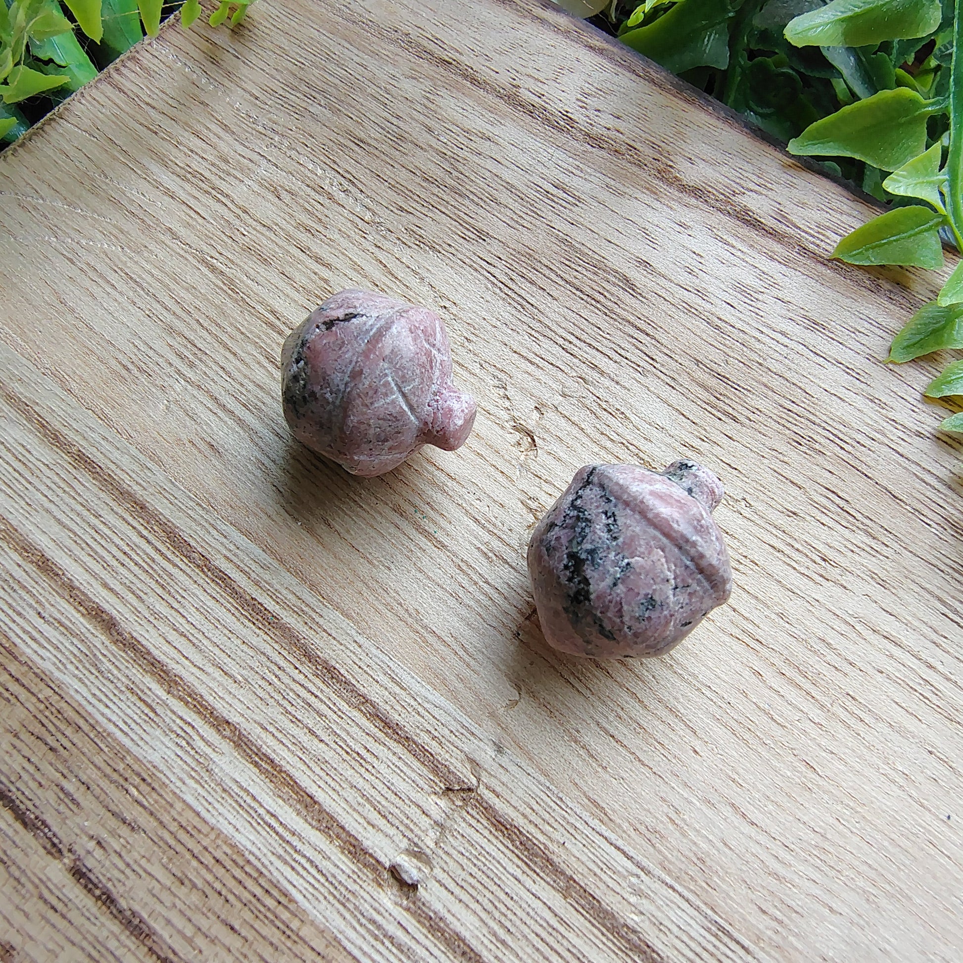 Two small rhodonite acorns on a wooden cutting board with greenery in the background